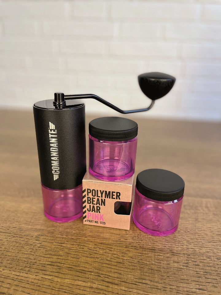 Comandante Bohnenbehälter in pink mit Deckel neben der Verpackung und einer Comandante Kaffeemühle